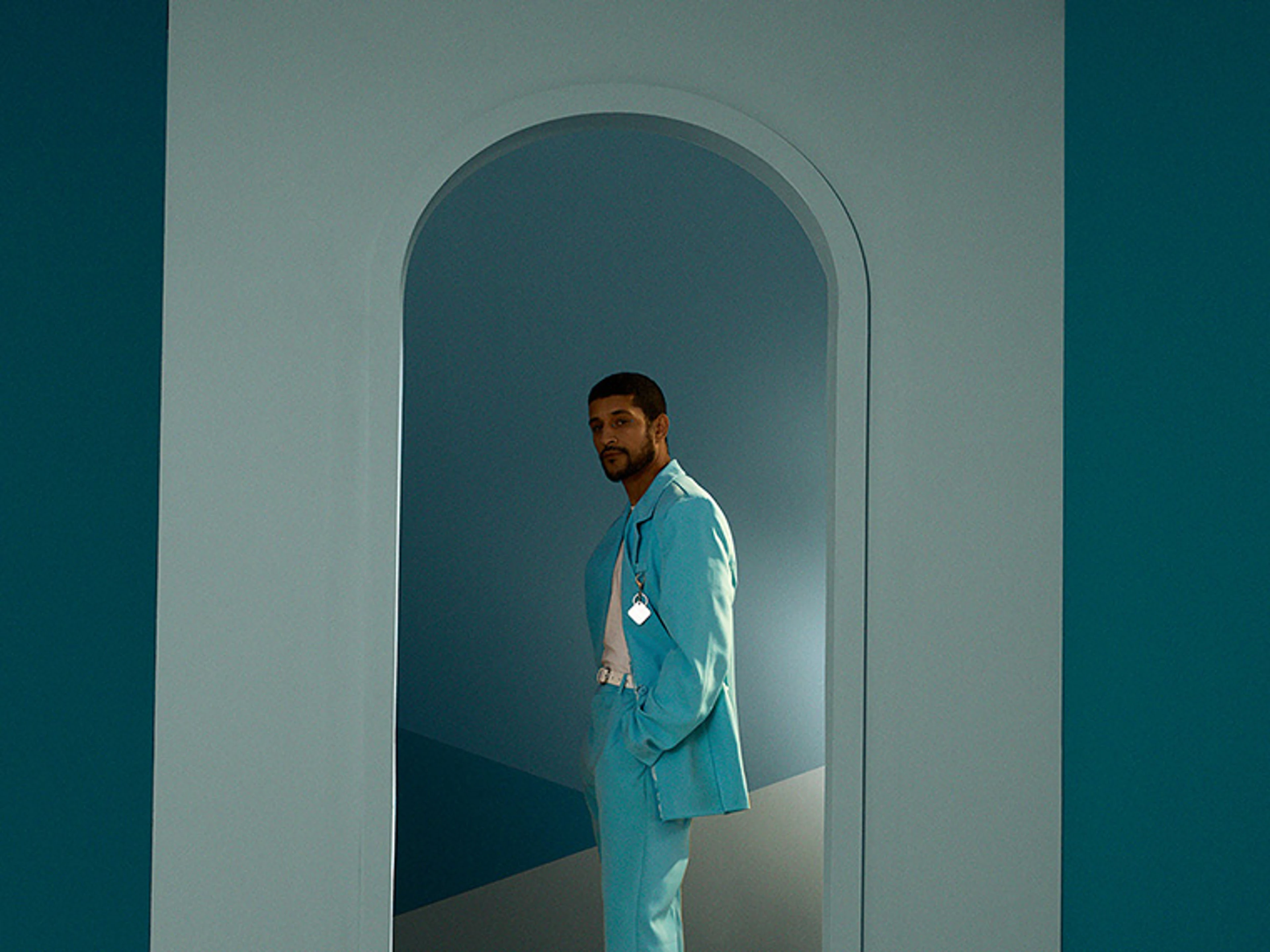 Person in a light blue suit stands in an arched doorway surrounded by walls in blue shades. The stylised interior highlights the interplay between architecture, light and colour.