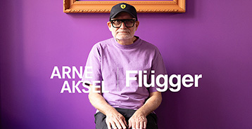 Stue med lilla vægge, hvor der sidder en hvidskægget mand. Manden har en lilla t-shirt, en sort kasket og sorte briller på.