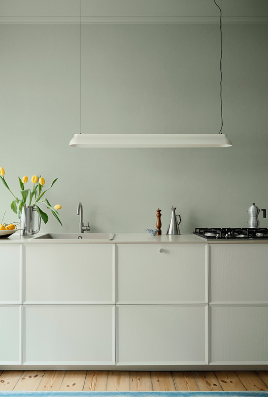 Køkken med lyse vægge, hvide elementer og træbordplade, indrettet med vask, komfur og aflang loftlampe.