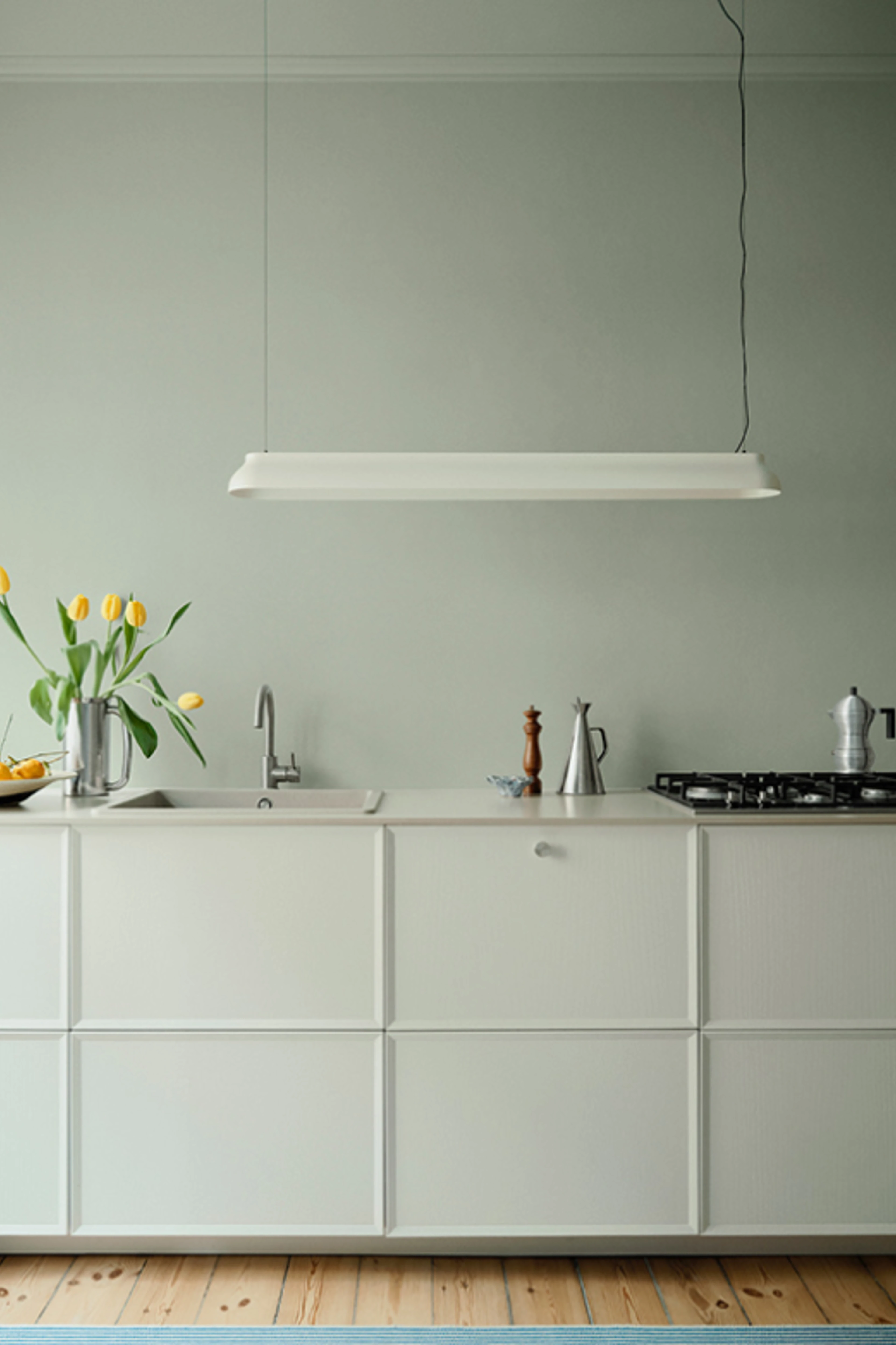 Køkken med lyse vægge, hvide elementer og træbordplade, indrettet med vask, komfur og aflang loftlampe.