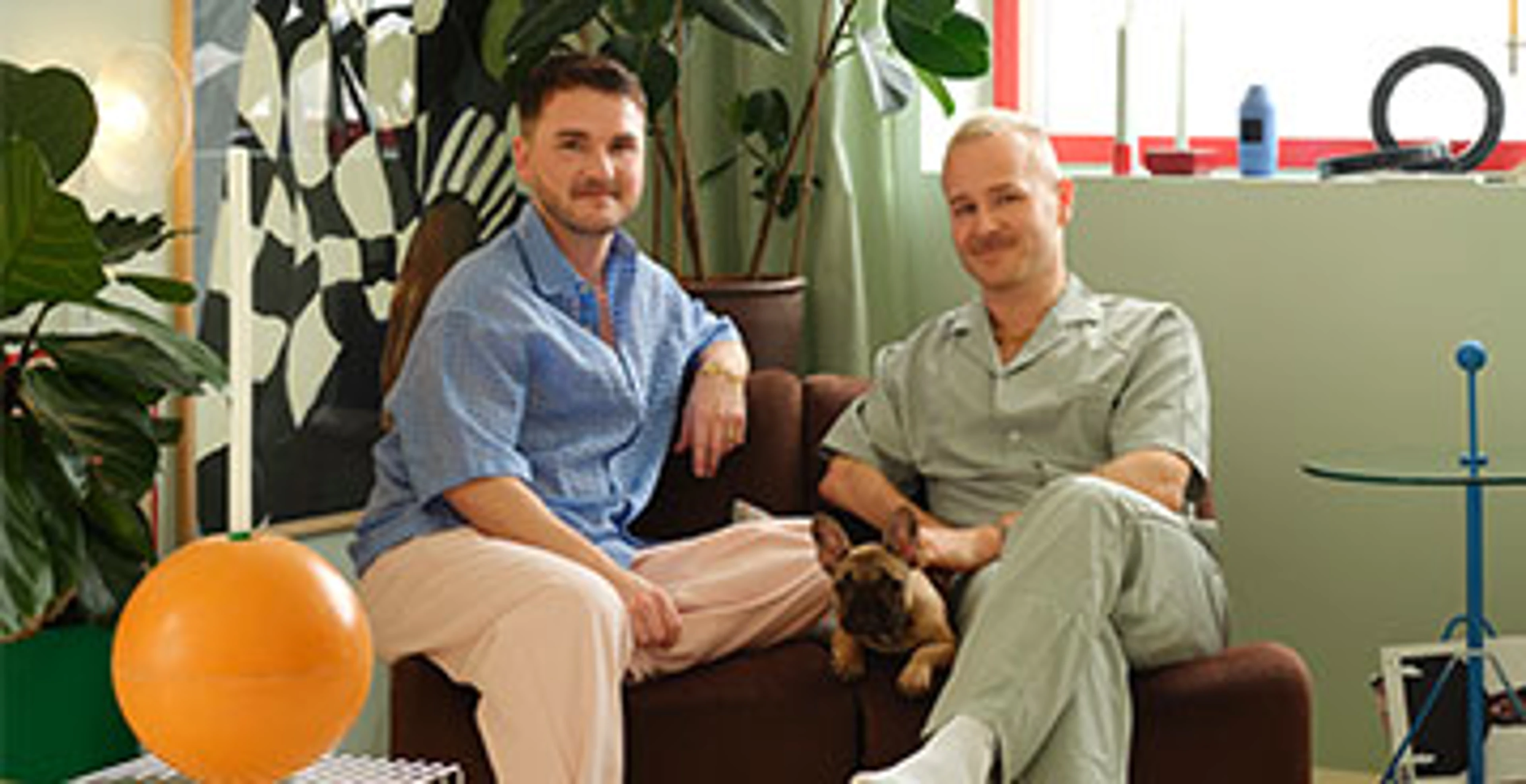 Two men sit together on a brown sofa with a small dog between them in a colorful living room.