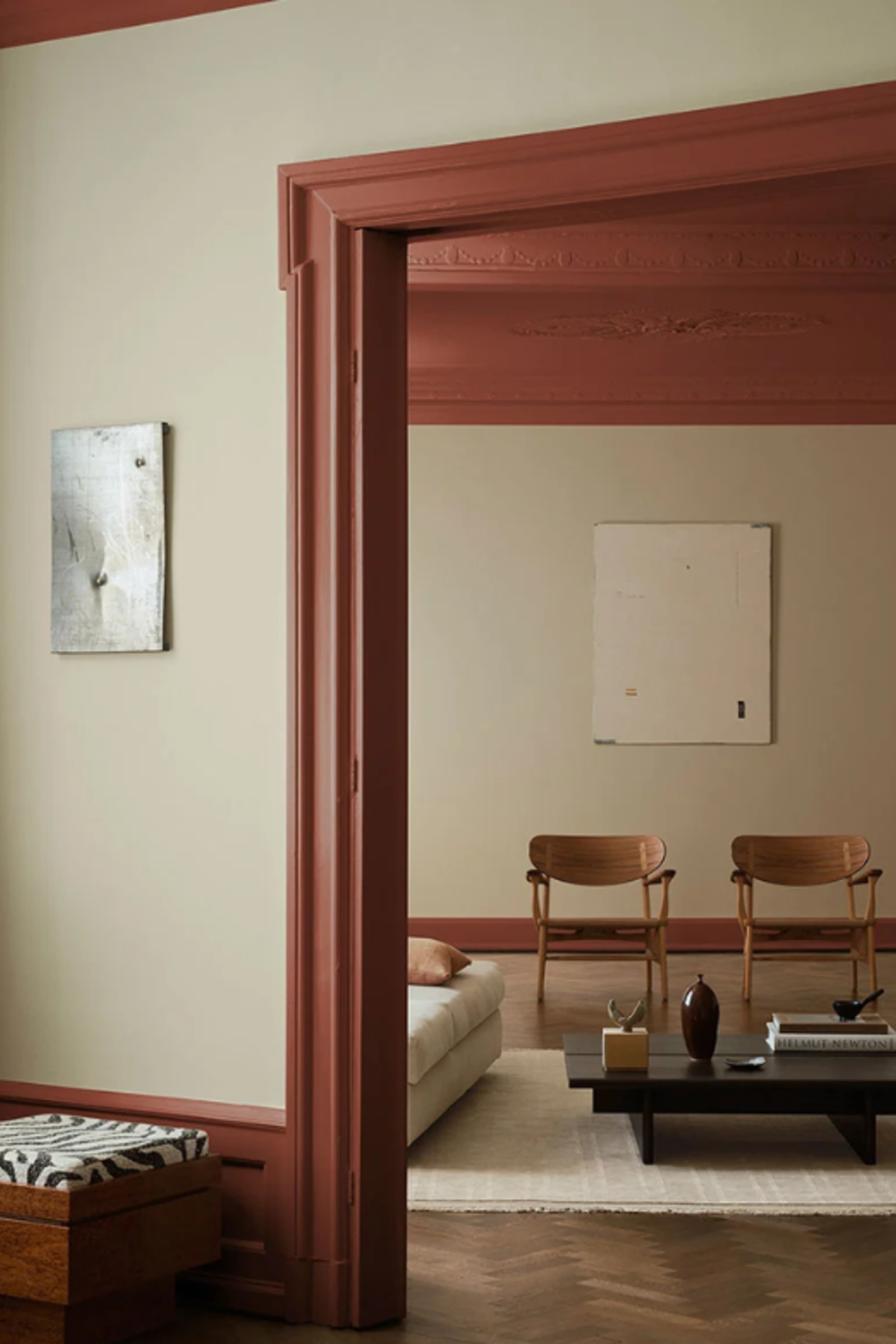 Rum med lys vægmaling og rødbrun vægmaling omkring døråbning, møbleret med lave stole, sofa og sofabord i et roligt, minimalistisk udtryk.