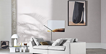 Stue med hvidlige vægge, en lysgrå sofa, og et sofabord med glasplade og marmorben. Der er en hvid lampe med skærm på sofabordet. På væggen er der er maleri.