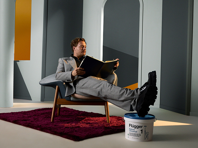 Person in a light grey suit sits relaxed in an armchair holding a large colour sample book. The room has geometric wall panels in grey and yellow shades, and a paint bucket with the Flügger logo stands in front of the chair.