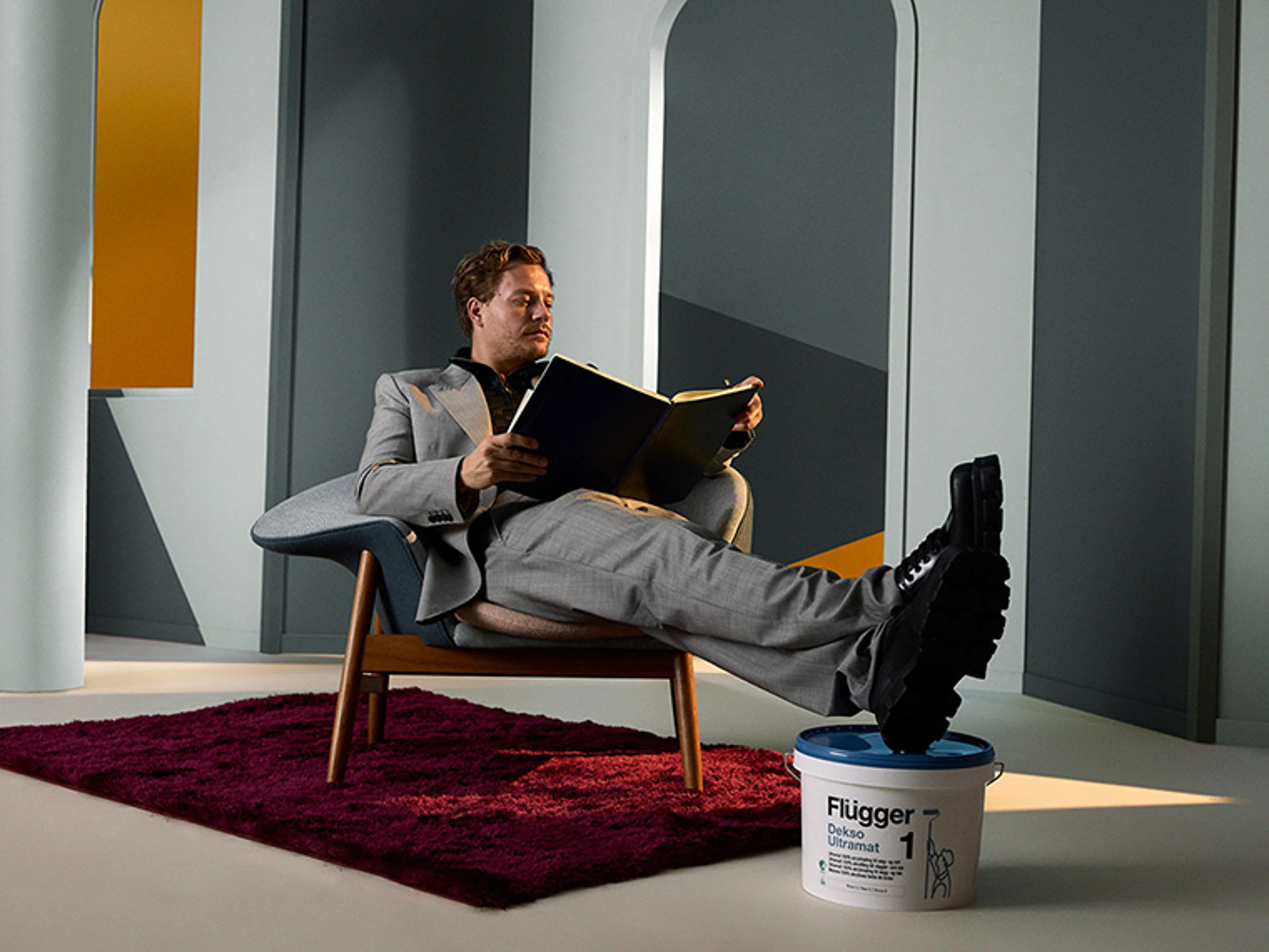 Person in a light grey suit sits relaxed in an armchair holding a large colour sample book. The room has geometric wall panels in grey and yellow shades, and a paint bucket with the Flügger logo stands in front of the chair.