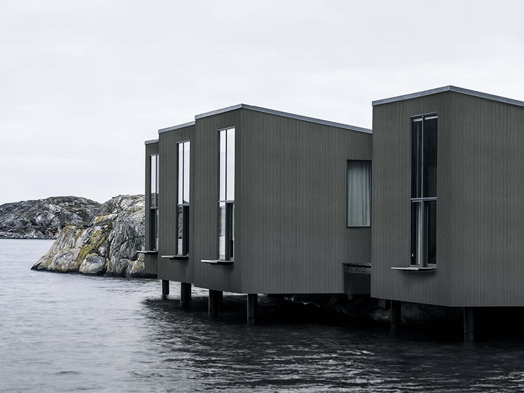 Moderna mörkgrå trähus på pålar över vattnet, med klippig kustlinje i bakgrunden.