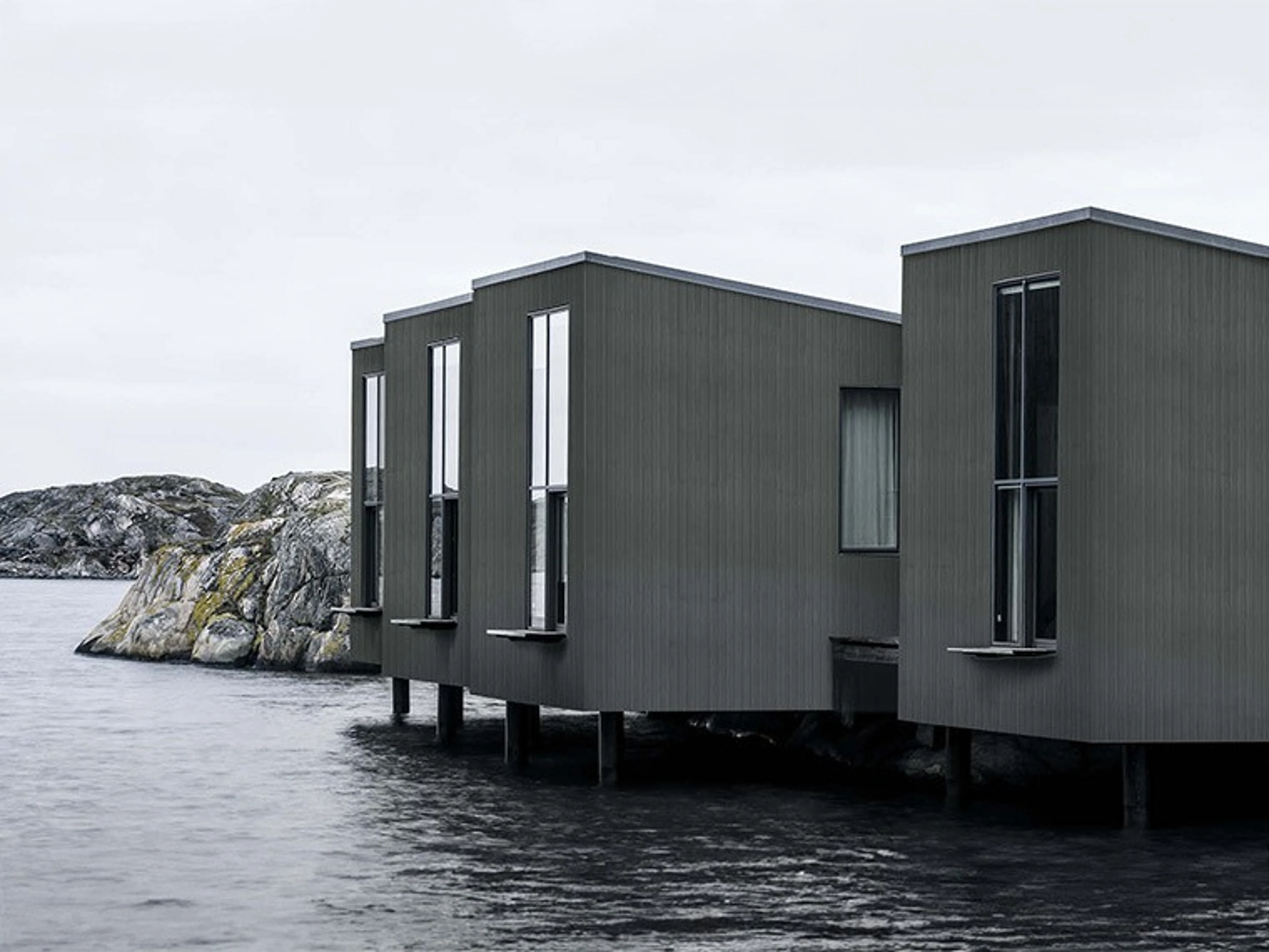 Moderna mörkgrå trähus på pålar över vattnet, med klippig kustlinje i bakgrunden.