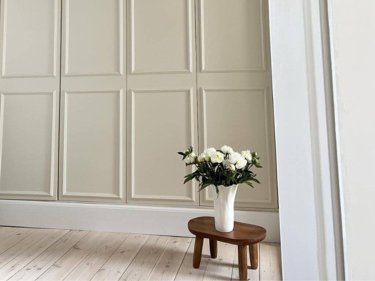 Beige skap og hvitpigmentert tregulv med en mørk trekrakk og en vase med hvite roser.