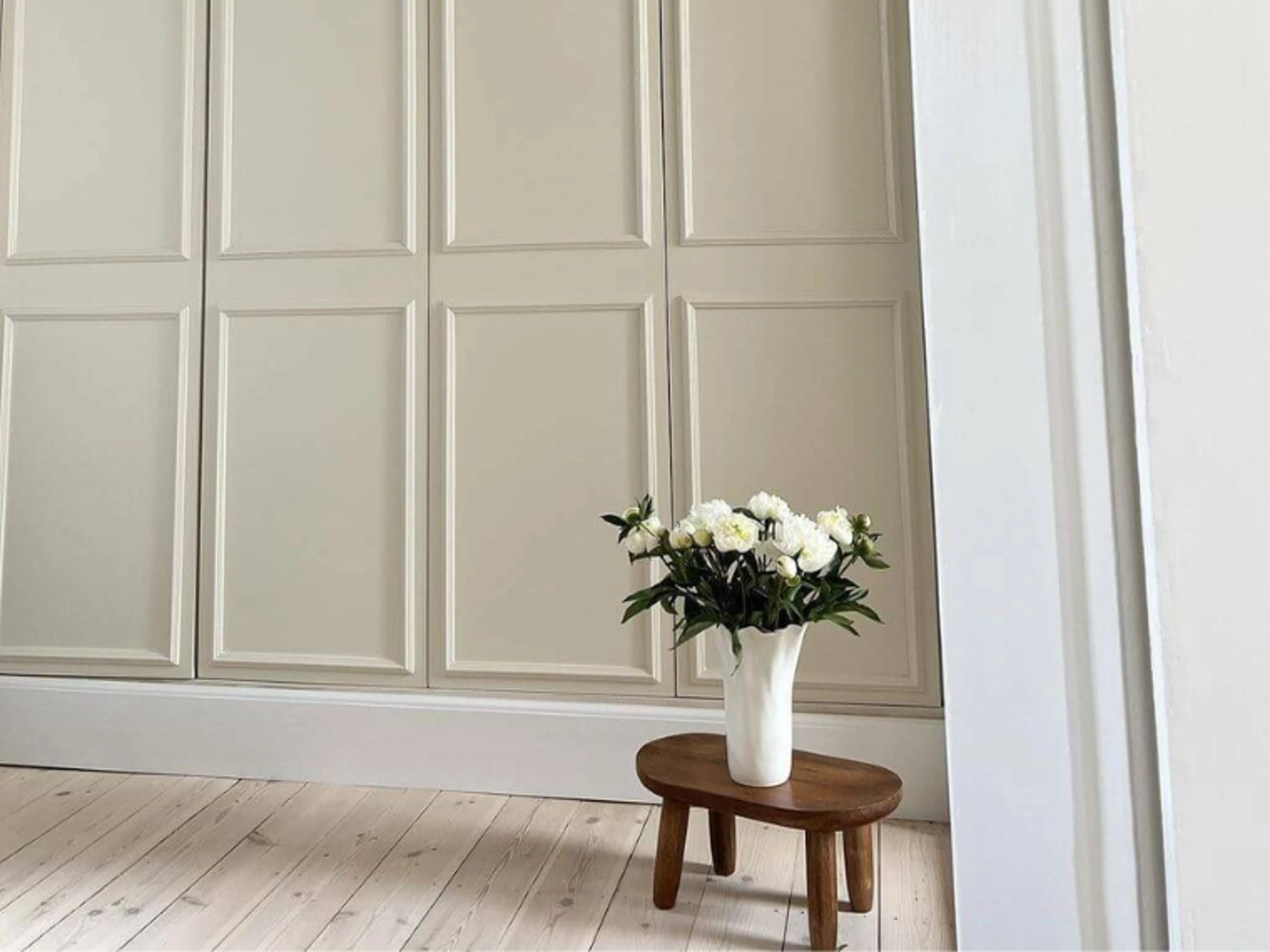 Beige skap og hvitpigmentert tregulv med en mørk trekrakk og en vase med hvite roser.