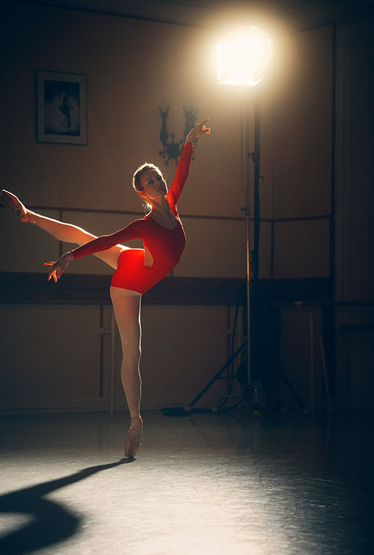 Balletdanser i rød dragt og hvide strømpebukser balancerer på tåspids i et mørkt studie oplyst af en kraftig lampe.