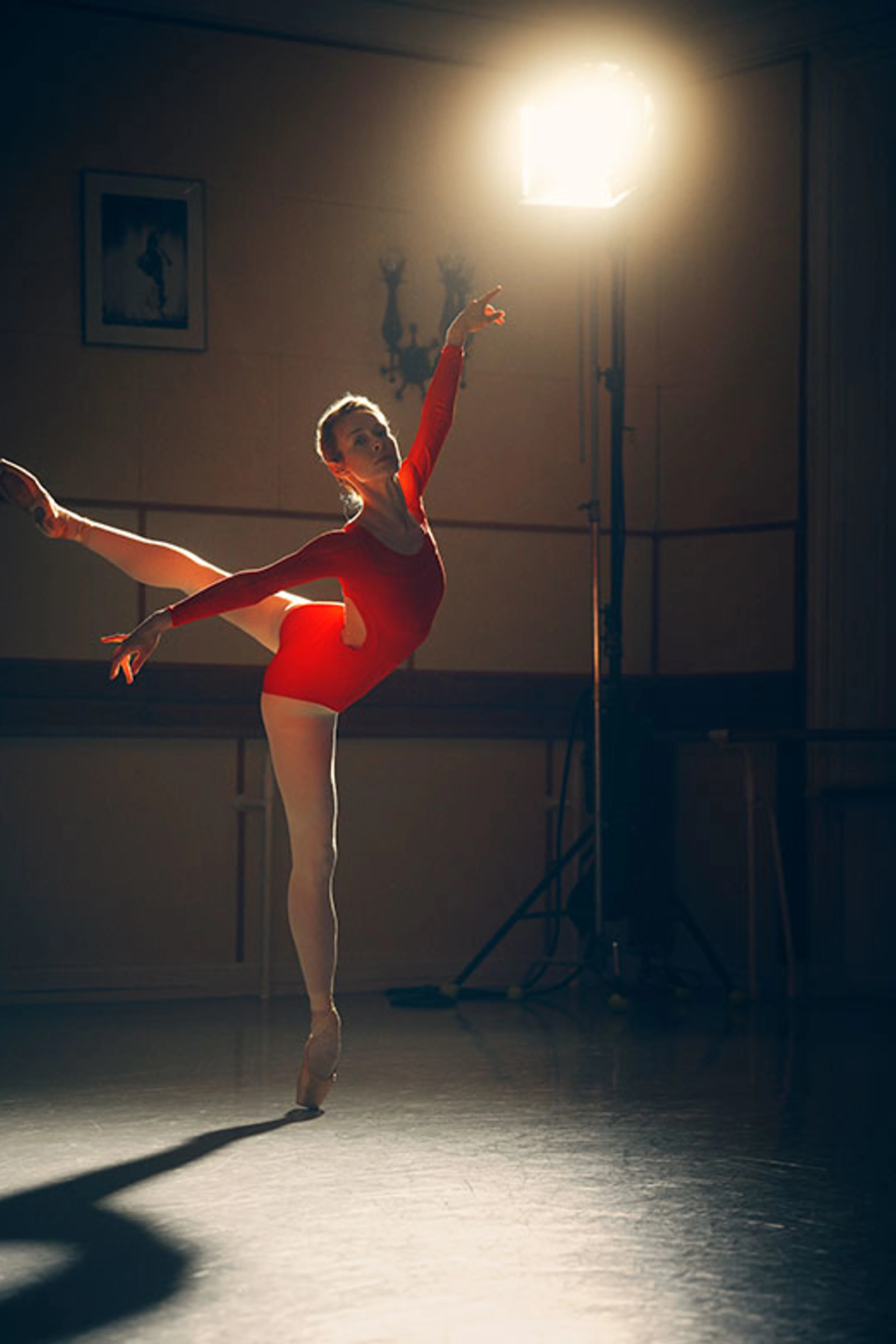 Balletdanser i rød dragt og hvide strømpebukser balancerer på tåspids i et mørkt studie oplyst af en kraftig lampe.