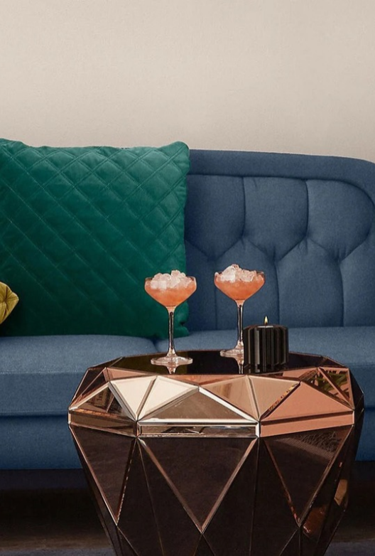 Blå sofa med knappheft og en grønn quiltet pute samt et geometrisk sofabord i bronsefarget speilglass med to cocktailer og et svart lys.