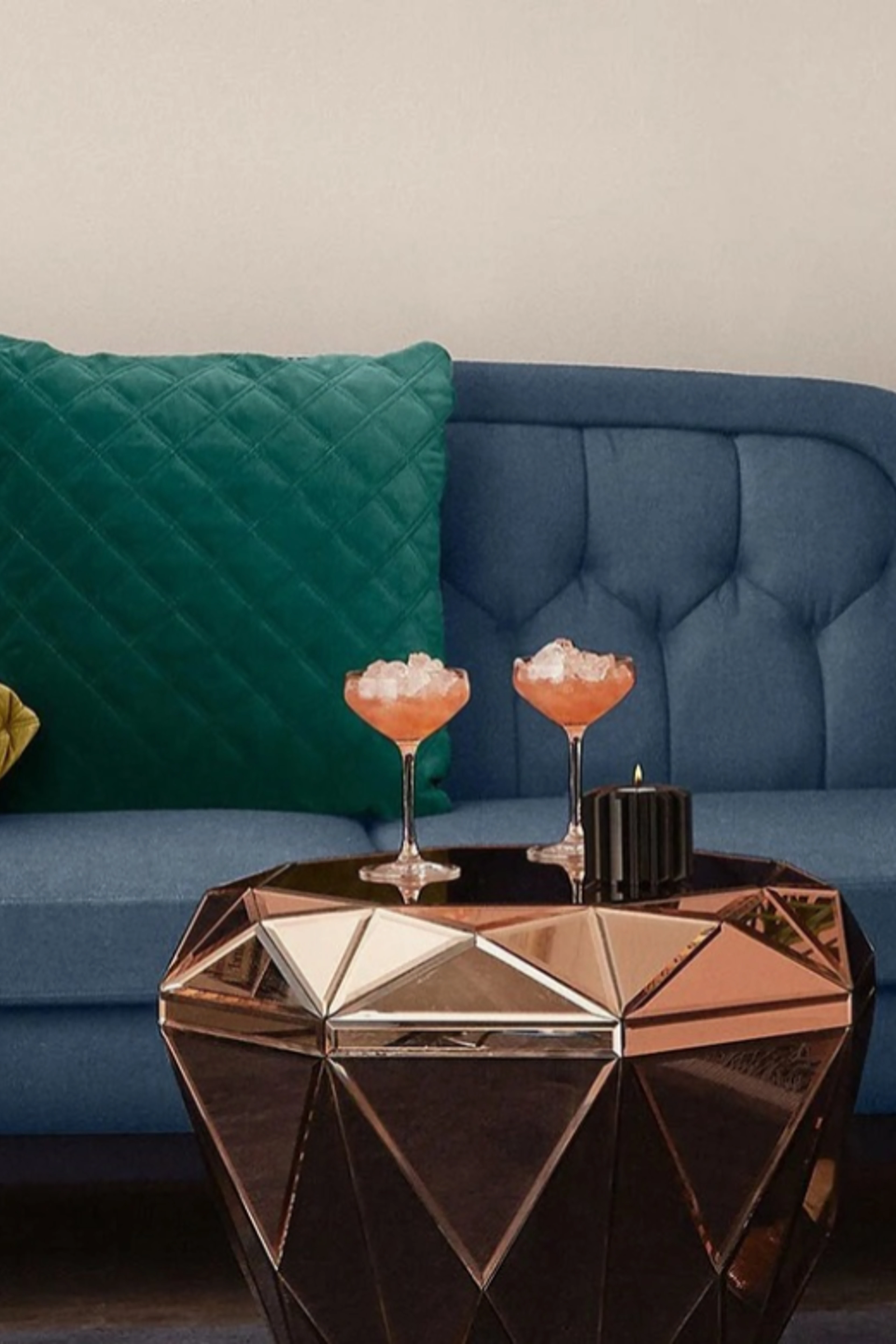 Blå sofa med knappheft og en grønn quiltet pute samt et geometrisk sofabord i bronsefarget speilglass med to cocktailer og et svart lys.