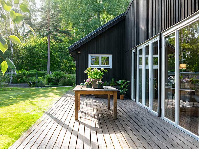 Sortmalet træhus med stor træterrasse og glaspartier ud mod en grøn have. På terrassen står et træbord med blomster.