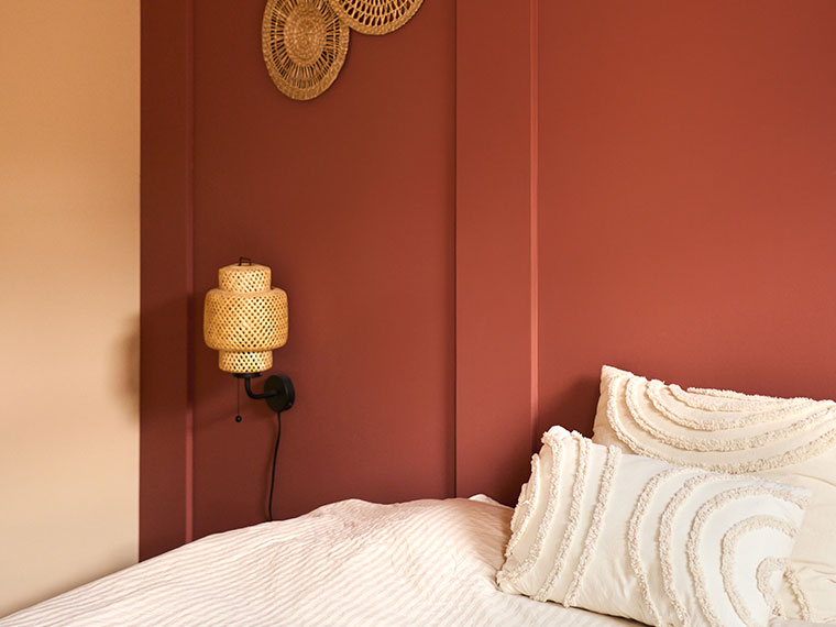 Bedroom with a deep terracotta-colored wall, light bedding, textured cushions, and a woven wall lamp.