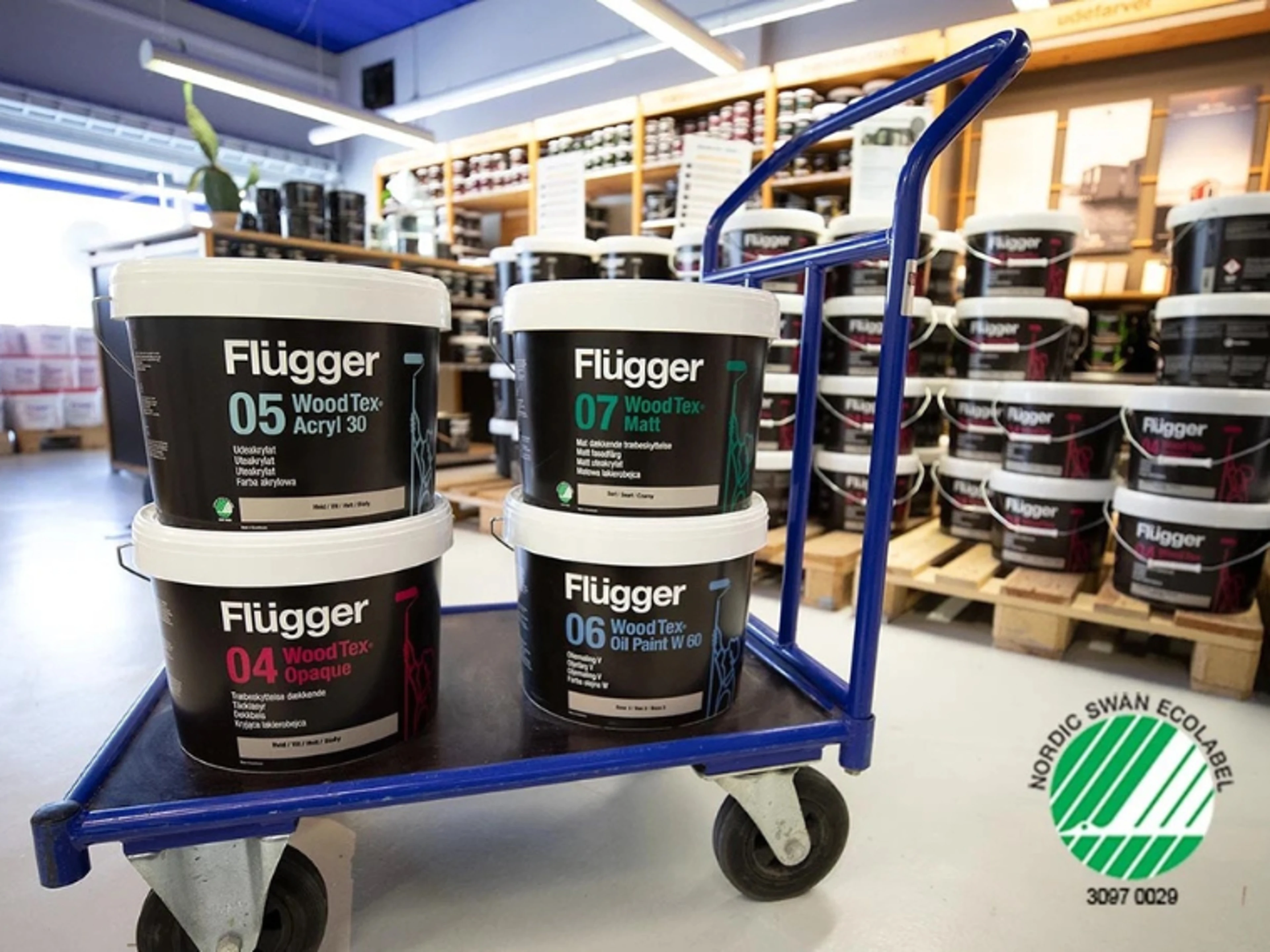 Fire spann Flügger WoodTex trebeskyttelse på en tralle i en butikk med hyller fulle av malingsspann i bakgrunnen, samt Svanemerket-logoen.