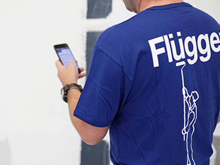 Person i blå Flügger-trøje bruger en smartphone foran en malet væg.