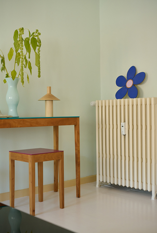 Bord og skammel i træ, indrettet med vase med grønne grene, lampe og radiator mod lys væg.