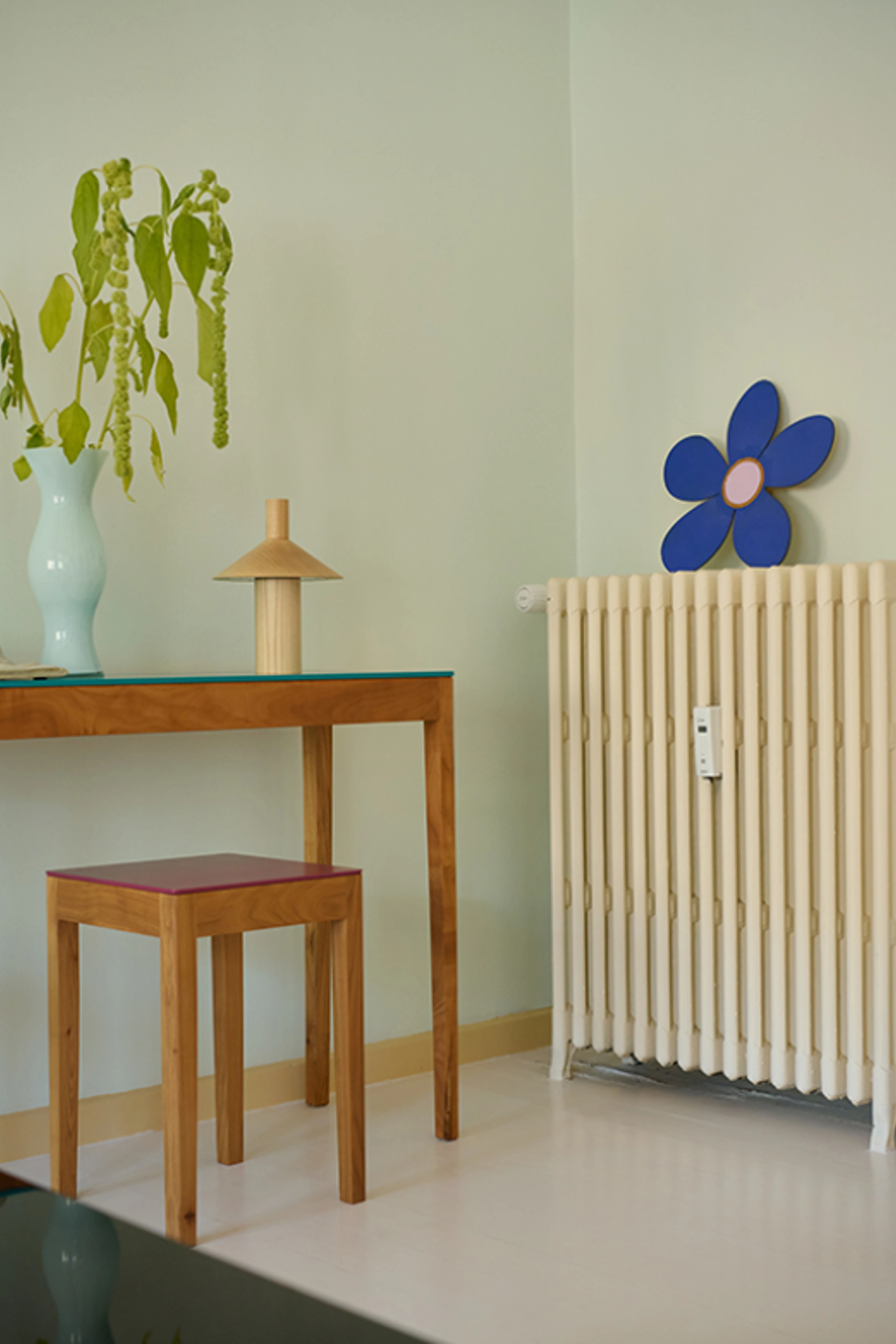 Bord og skammel i træ, indrettet med vase med grønne grene, lampe og radiator mod lys væg.