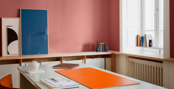 Kontor med rosamalt vegg, hvitt skrivebord med oransje skriveunderlag, et blått innrammet bilde og en oransje stol foran et høyt vindu.