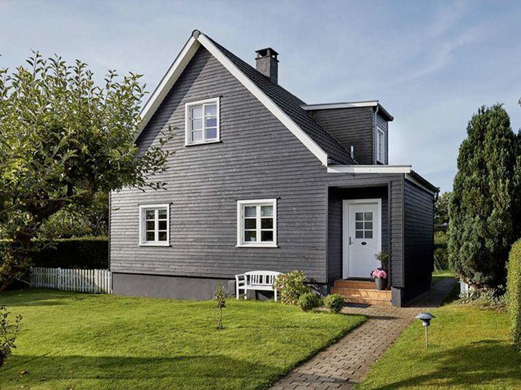 Villa med mørk fasade og hvite vindusrammer, omgitt av en velstelt hage. En sti fører gjennom plenen frem til husets inngang.
