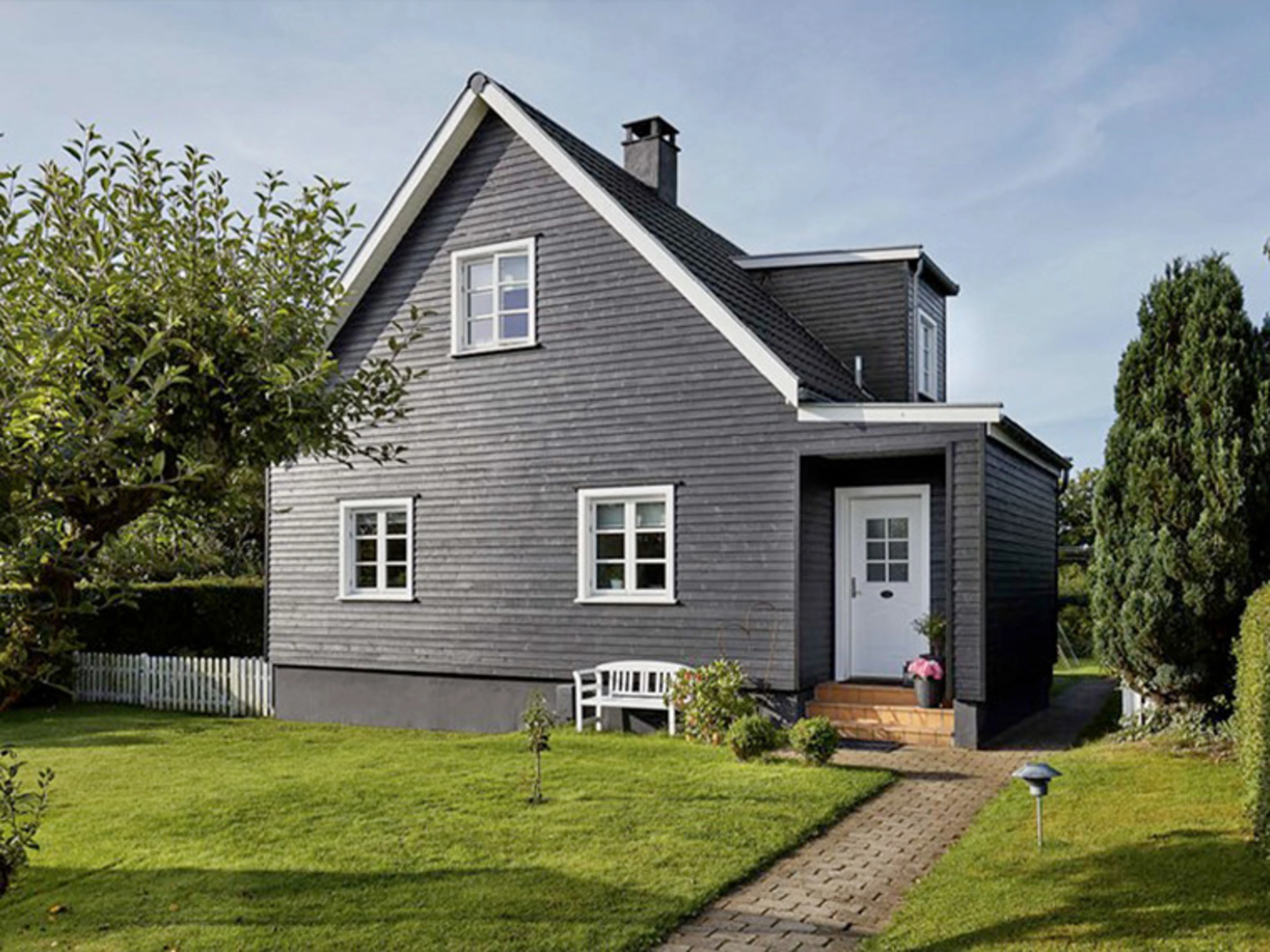 Villa med mörk fasad och vita fönsterkarmar, omgiven av en välskött trädgård. En stig leder genom gräsmattan fram till husets entré.