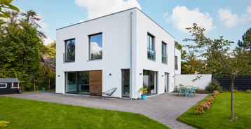Moderne hvitt hus i to etasjer med store vinduer, steinlagt terrasse og grønn hage.