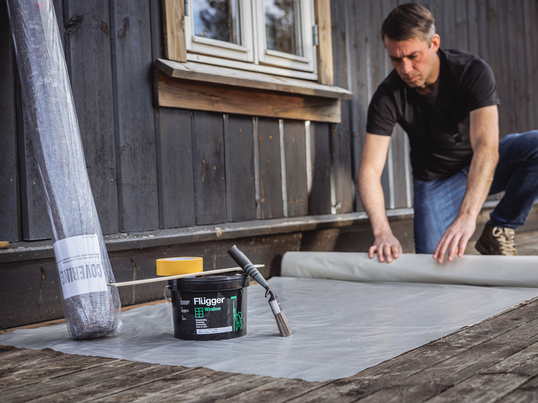 A man is rolling out protective plastic sheeting on the terrace while preparing to paint the windows, with a bucket of Flügger Window paint, a paintbrush, painter’s tape, a stir stick, and a roll of protective felt nearby.