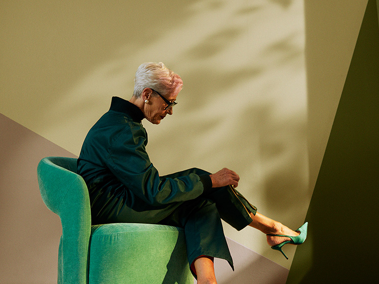 Person sits in a green armchair placed in a stylised interior with geometric walls in muted green and beige shades. The image highlights the interplay between furniture, lighting and wall colours in a modern interior setting.