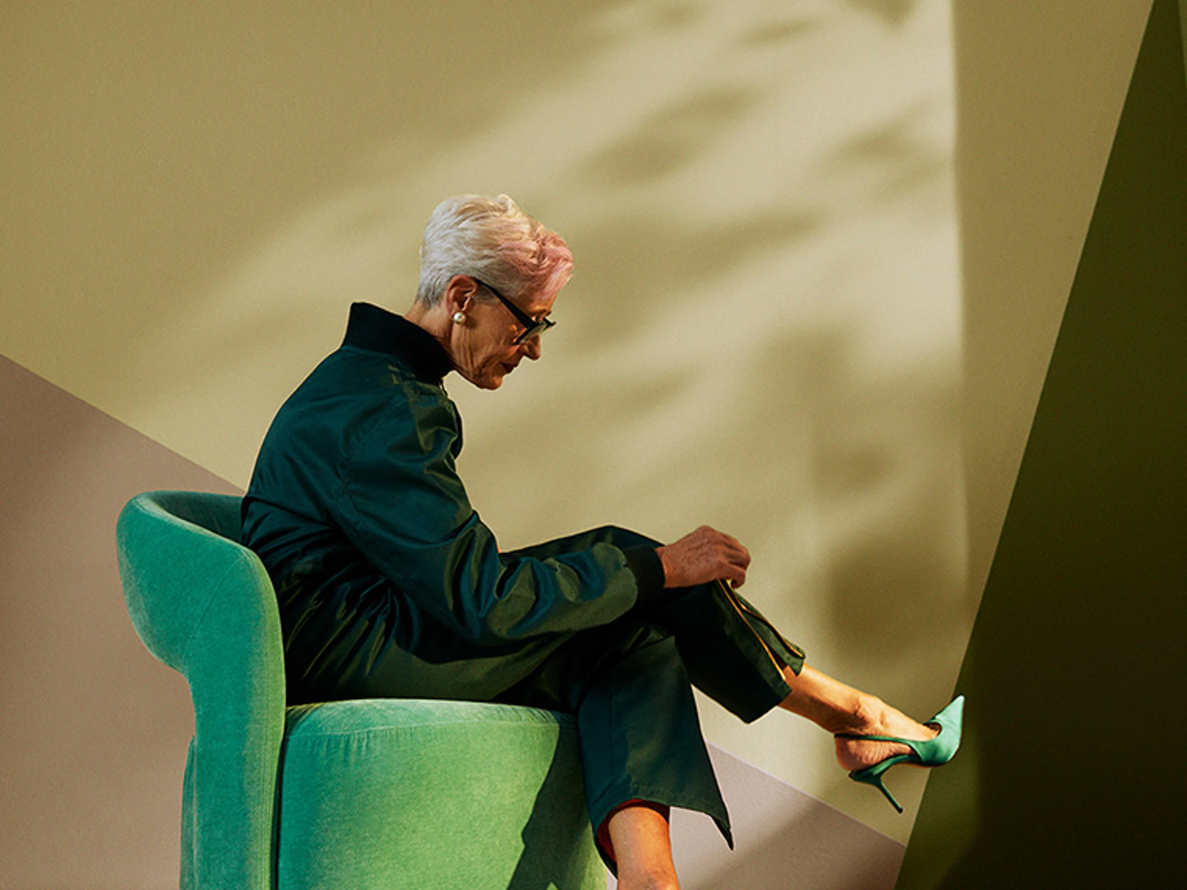 Person sits in a green armchair placed in a stylised interior with geometric walls in muted green and beige shades. The image highlights the interplay between furniture, lighting and wall colours in a modern interior setting.
