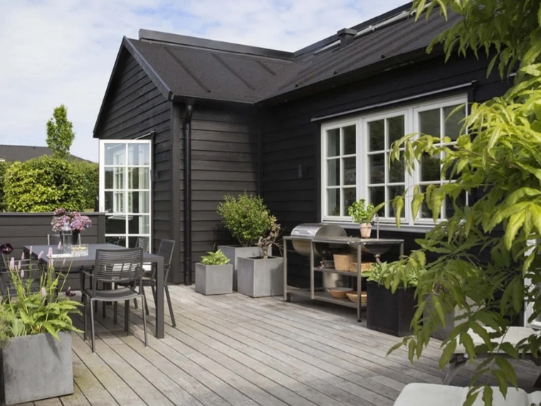 Wooden holiday home with black wood protection paint and a wooden terrace with many plants.