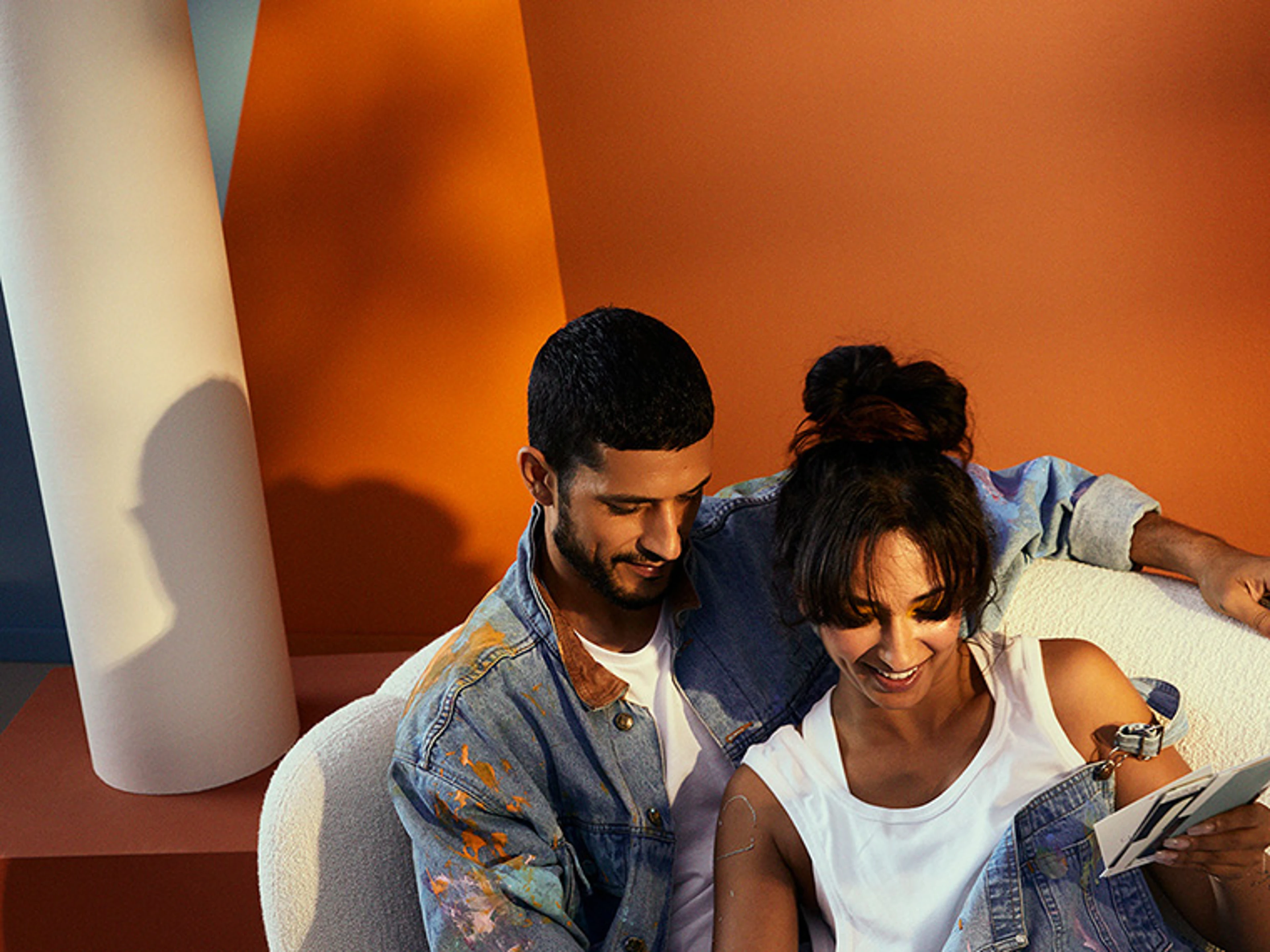 Two people sit close together in a white upholstered chair in front of walls with geometric colour blocks in orange, blue and neutral shades. One person holds a series of colour samples, while the interior highlights modern colour combinations and design.
