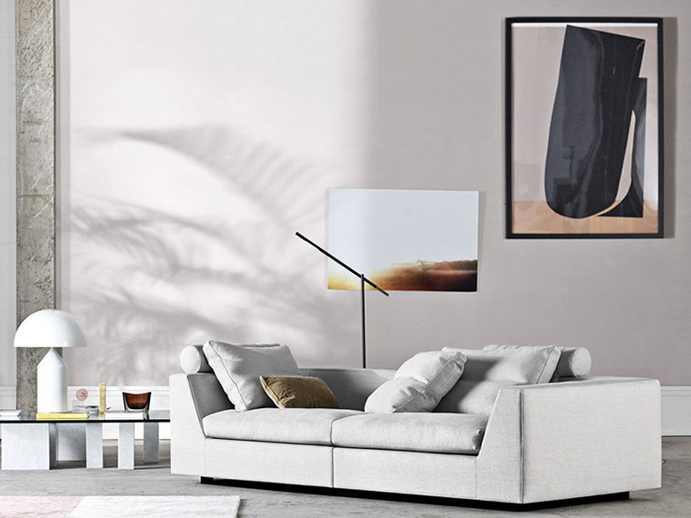 Stue med hvitlige vegger, en lysegrå sofa og et sofabord med glassplate og marmorben. Det er en hvit lampe med skjerm på sofabordet. På veggen er det et maleri.