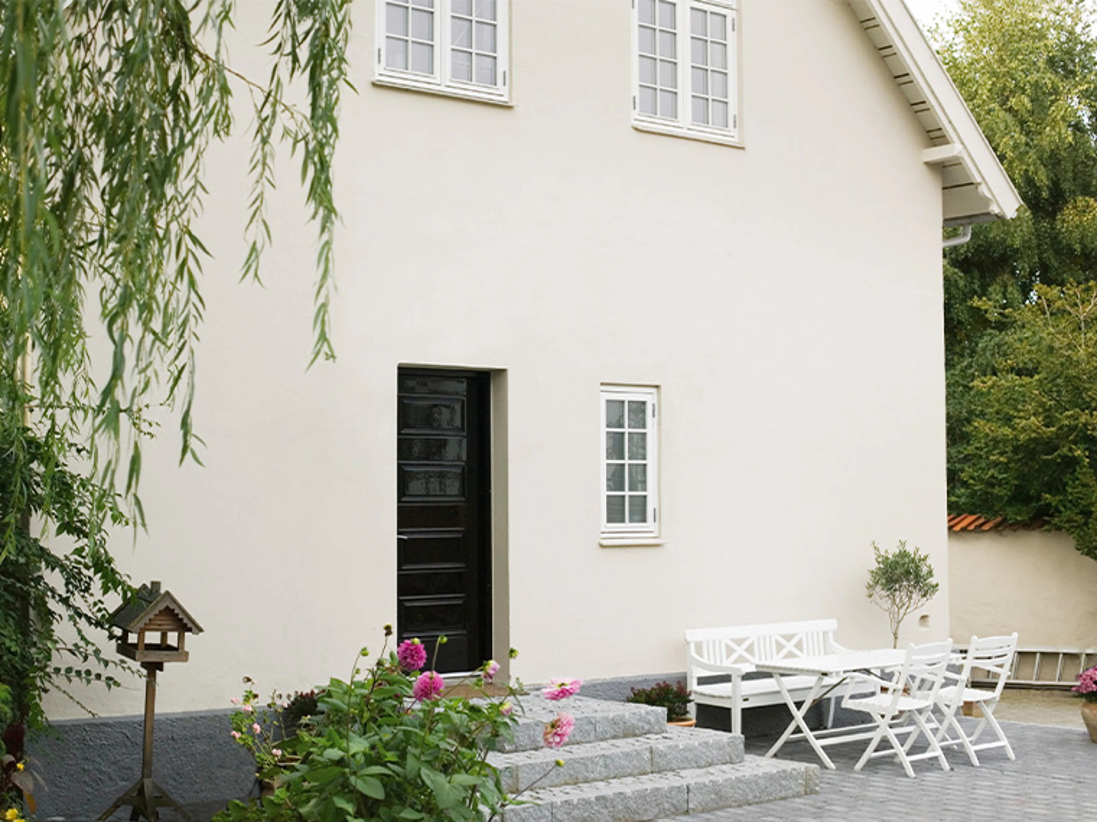 Hvitt hus med svart ytterdør, hvitinnrammede vinduer og terrasse med hvite hagemøbler.