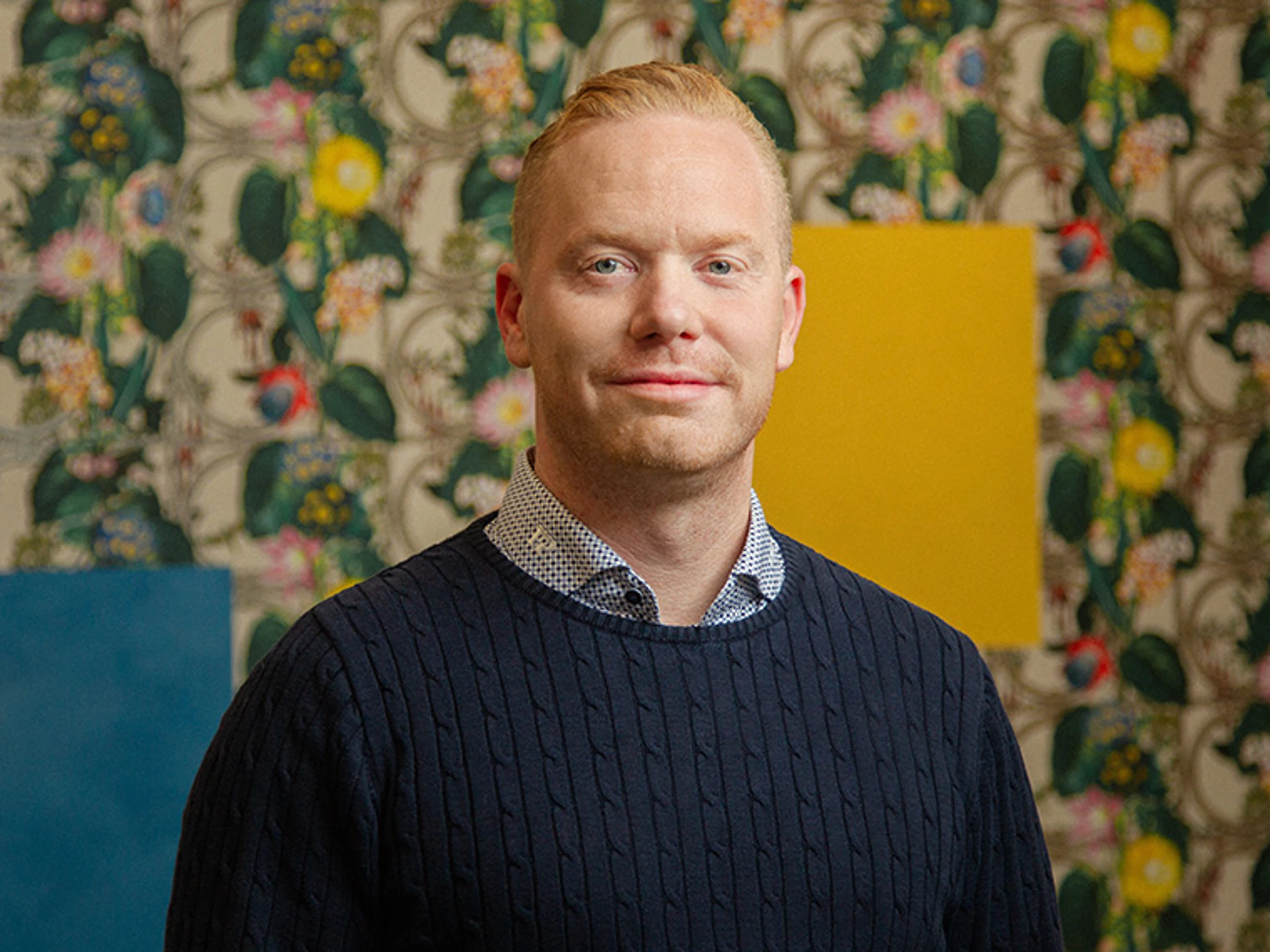 Mikkel Boelskite foran en væg med blomstrede tapetmønstre. I baggrunden ses farvede prøveplader i blå og gul, som fremhæver indretningens farver og materialer.