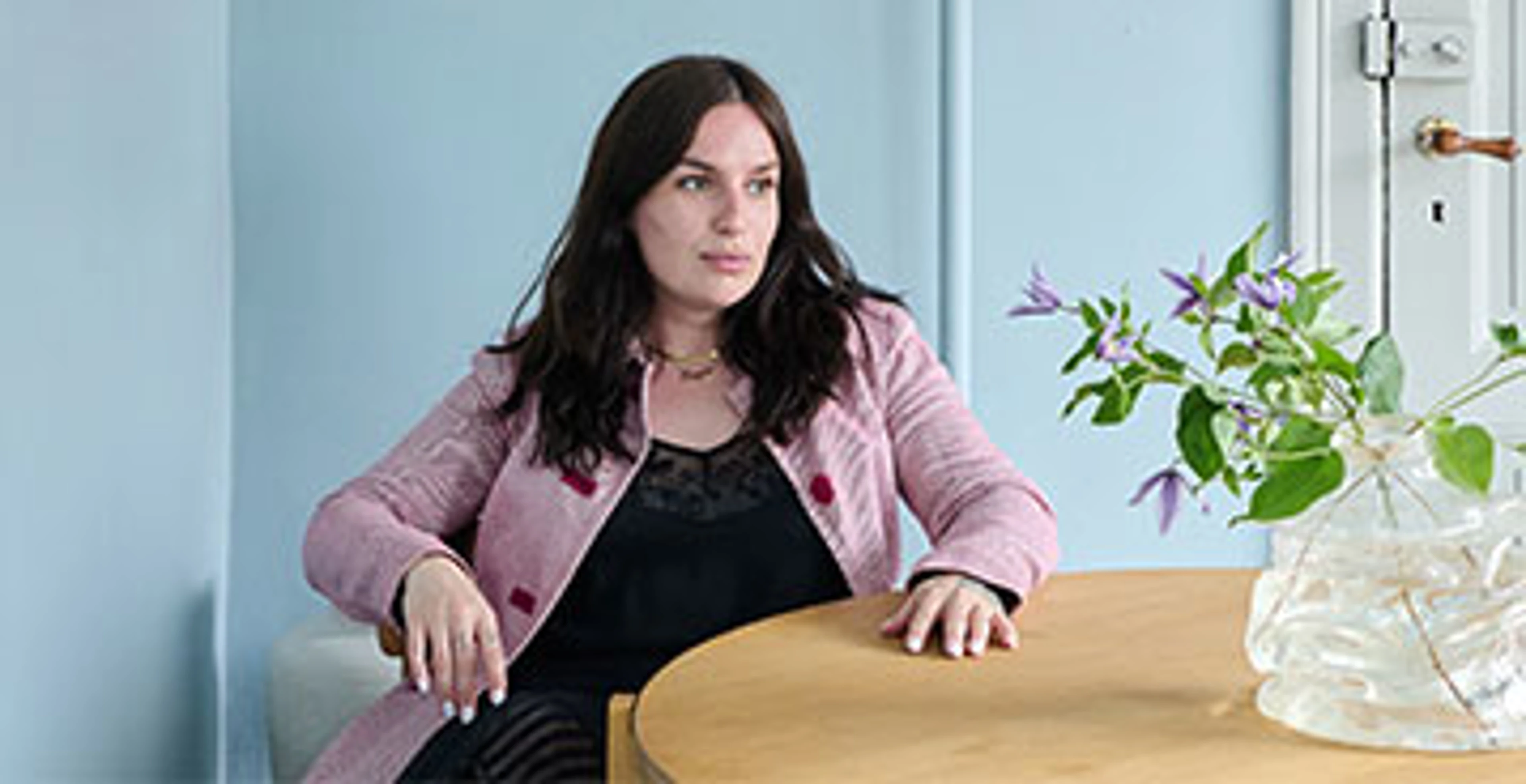 Person siddende ved rundt spisebord med lyseblå vægge, blomster i glasvase og dagslys fra vinduet.