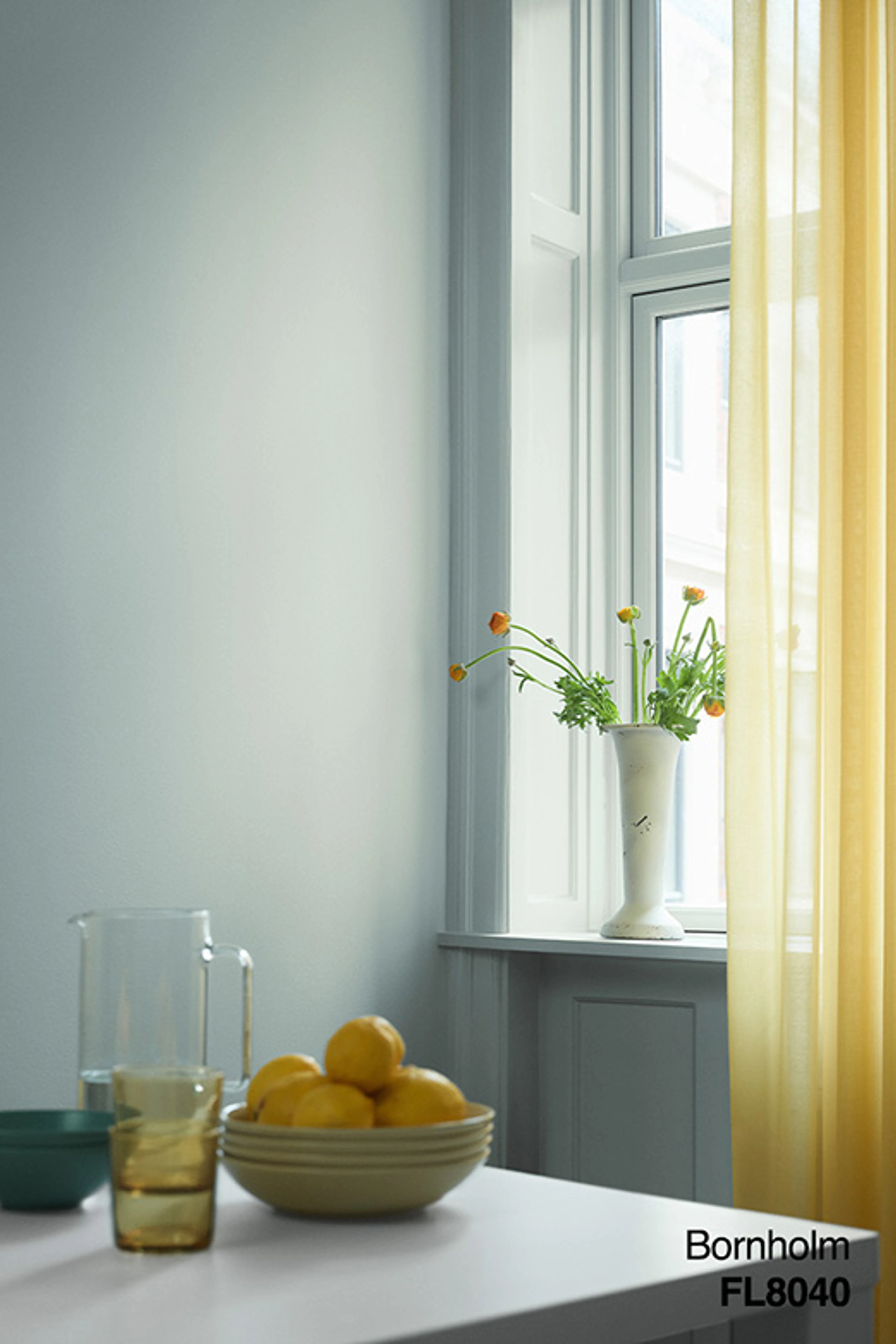 Ljust matplats med ljusgrå väggar, gula genomskinliga gardiner, vitt fönster och bord med citroner, glas och vas med blommor.