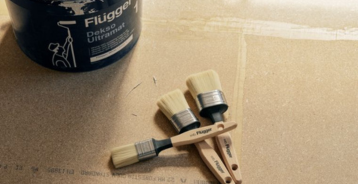 A bucket of Flügger Dekso Ultramat paint next to three paintbrushes, placed on a sheet of plywood.