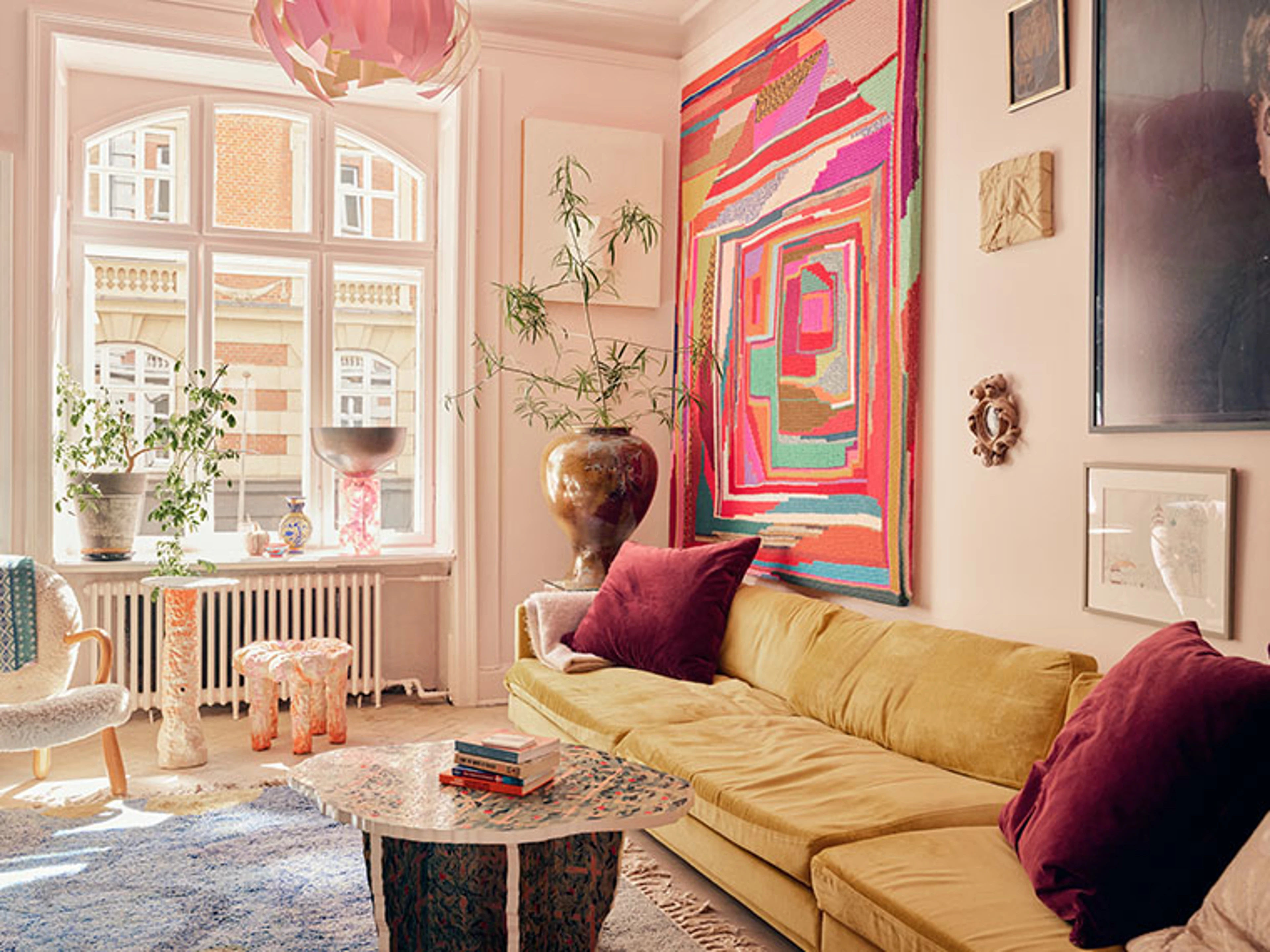 Farverig stue med gul sofa, stort abstrakt vægtæppe, kunst på væggene og planter ved et højt vindue.