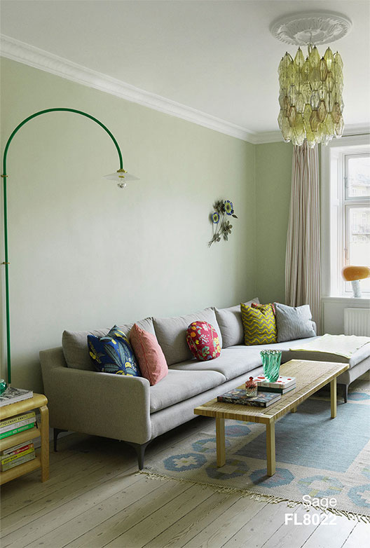 Stue med vægge i Sage, indrettet med lys sofa, farverige puder, træbord og grøn væglampe samt glaslysekrone.