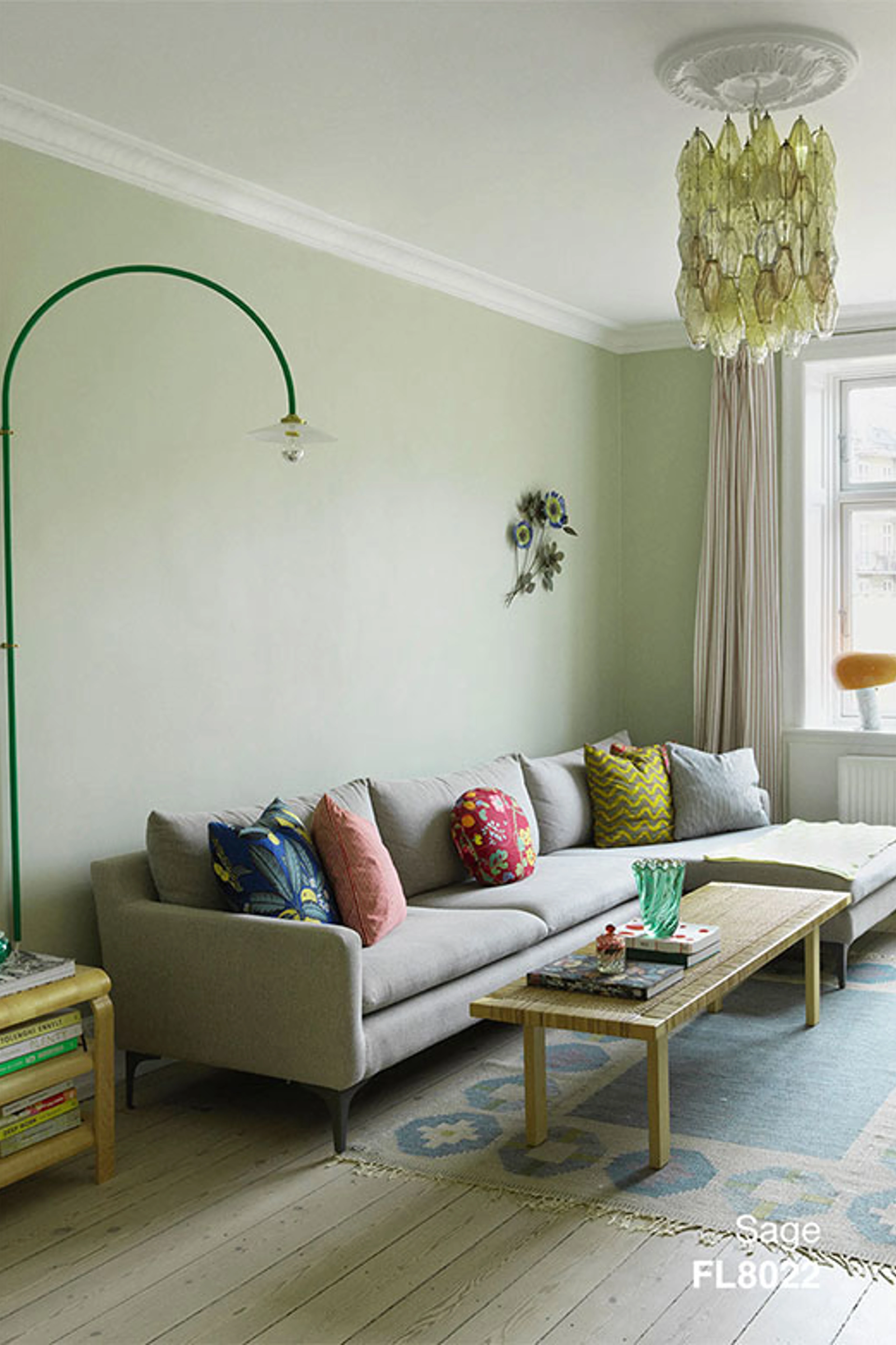 Stue med vægge i Sage, indrettet med lys sofa, farverige puder, træbord og grøn væglampe samt glaslysekrone.
