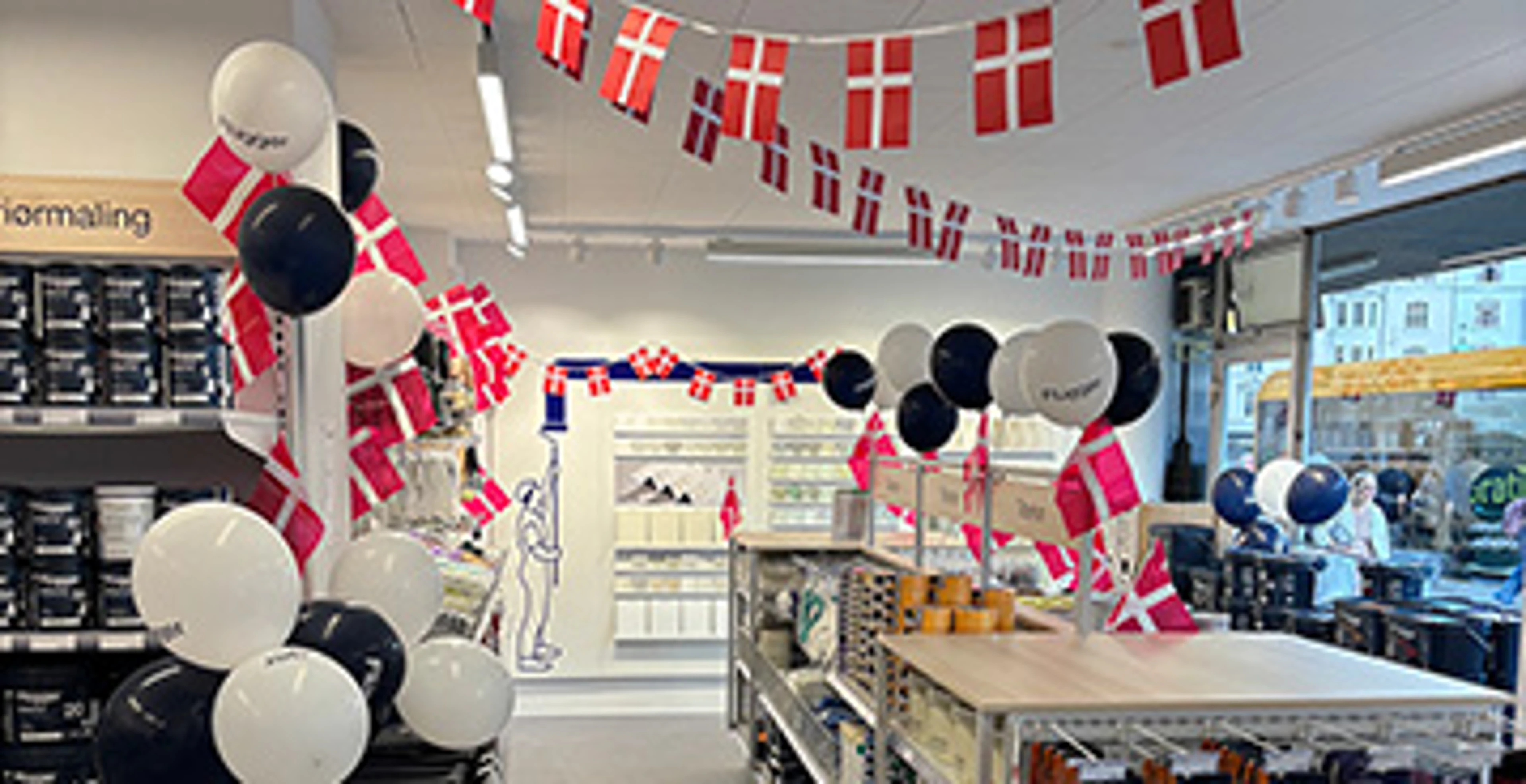 Interiør fra en Flügger-butik dekoreret med danske flag og balloner i rød, hvid og mørkeblå. Butikken indeholder hylder med maling, malertilbehør og udstillingsområder, mens vinduerne ud mod gaden lukker dagslys ind.