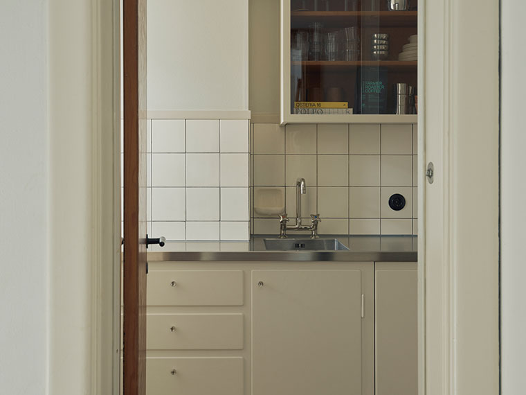 View through a doorway into a compact kitchen with cream-colored cabinets, white tiles, a stainless steel sink, and a glass-fronted cabinet.