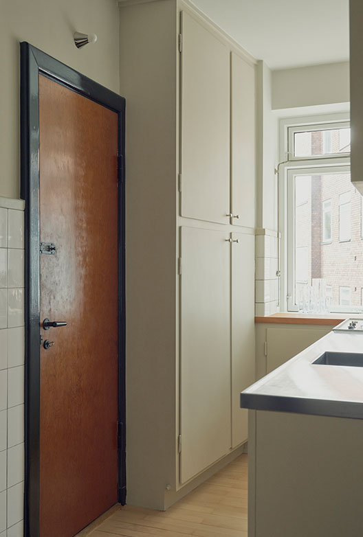 Narrow, bright kitchen with tall cream-colored cabinets, a wooden door, stainless steel worktable, and a large window.