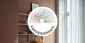 Farvegaranti-logo placeret foran et interiør med brun væg, åben reol og vindue. Logoet viser en vifte af farveprøver i forskellige nuancer.