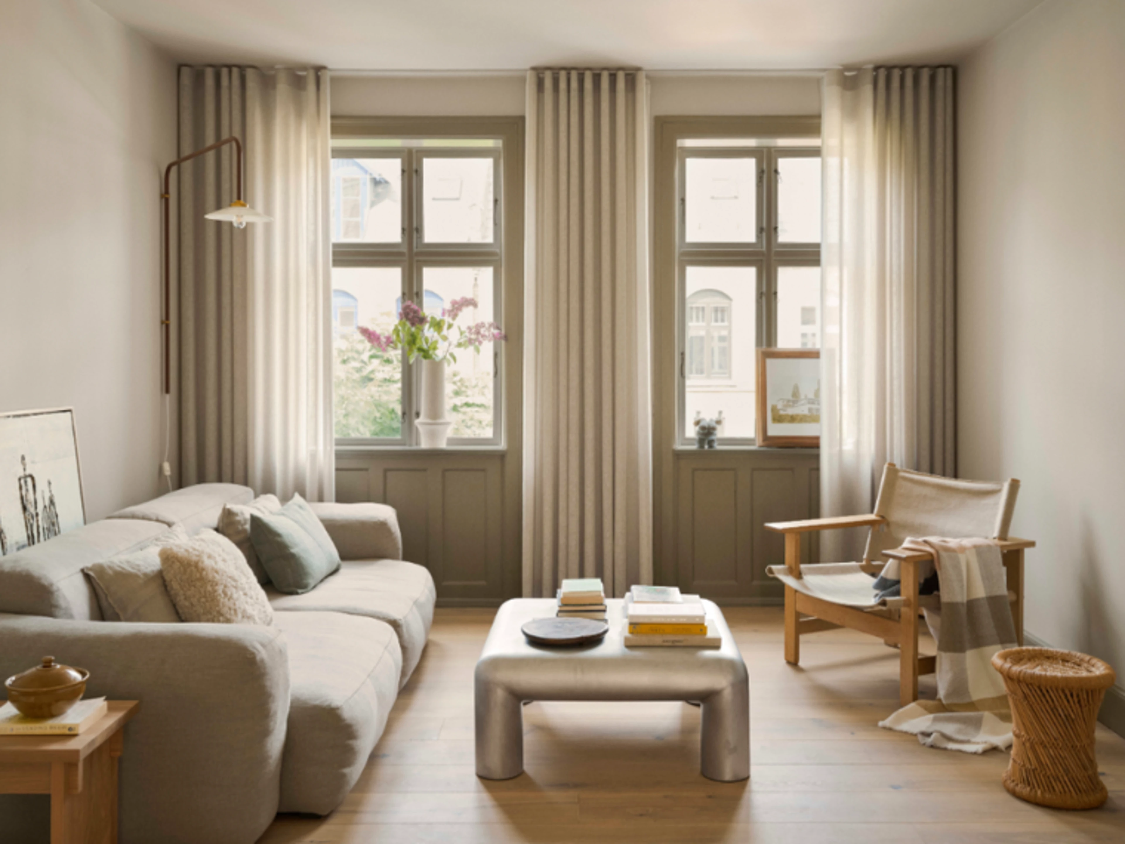 Stue med en lys sofa, sofabord og lænestol foran en varm beige væg. Store vinduer med lyse gardiner lukker dagslys ind, og der står blomster i vindueskarmen.