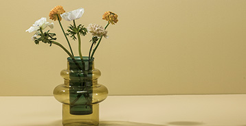 Grøn glasvase med hvide og gule blomster placeret foran en varm gul væg i en enkel og dekorativ opstilling.