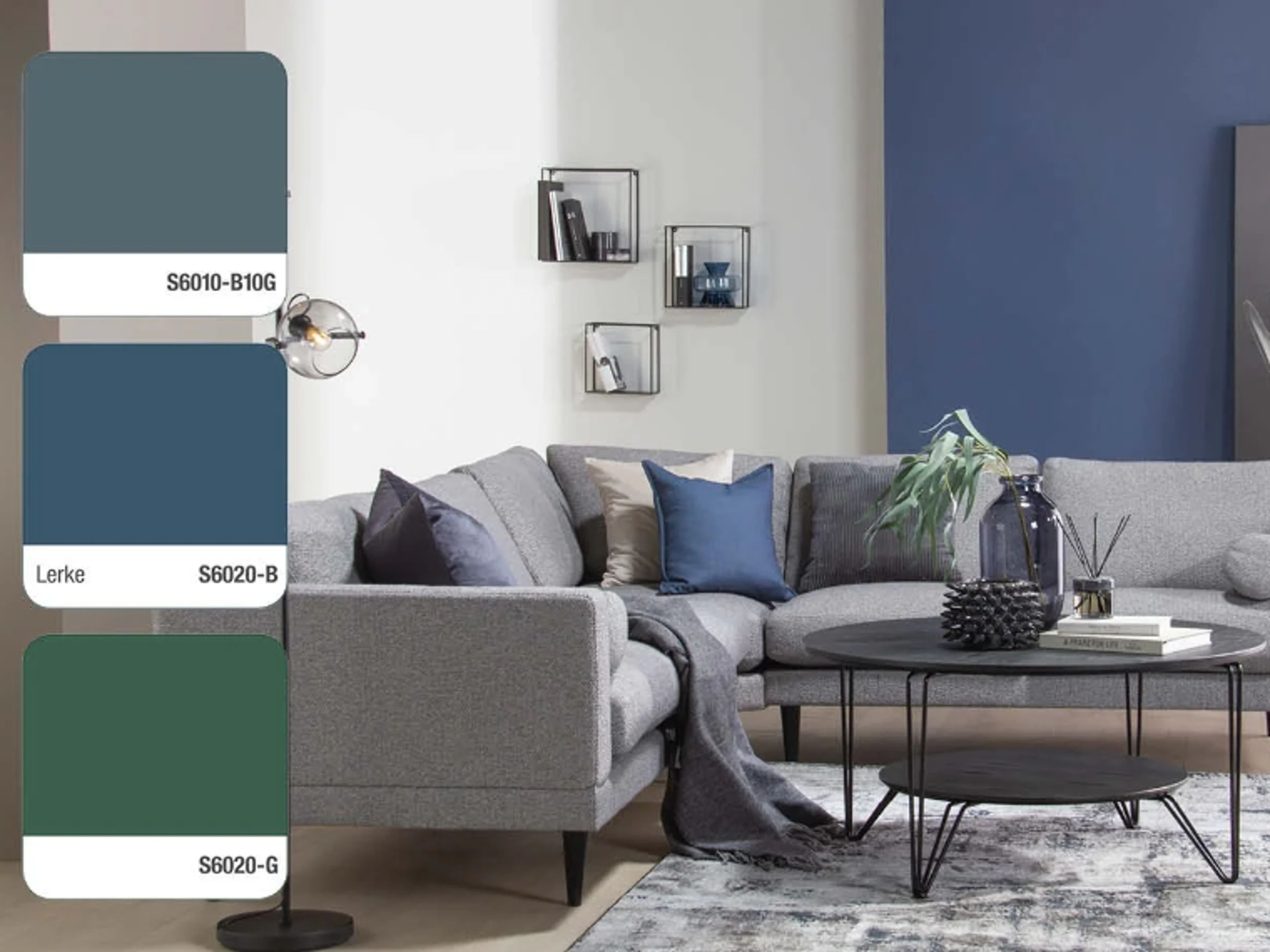 Living room with white and blue walls, a light grey corner sofa with blue and grey cushions, a round black coffee table, wall-mounted shelf cubes and a glass globe lamp, with three colour swatches on the left: S6010-B10G, Lerke S6020-B and S6020-G.
