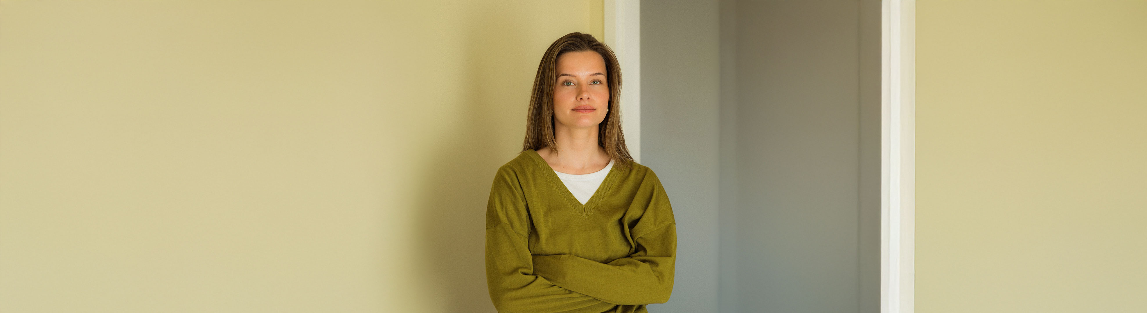 Woman with long brown hair in an olive-green sweater in front of a pale yellow wall.