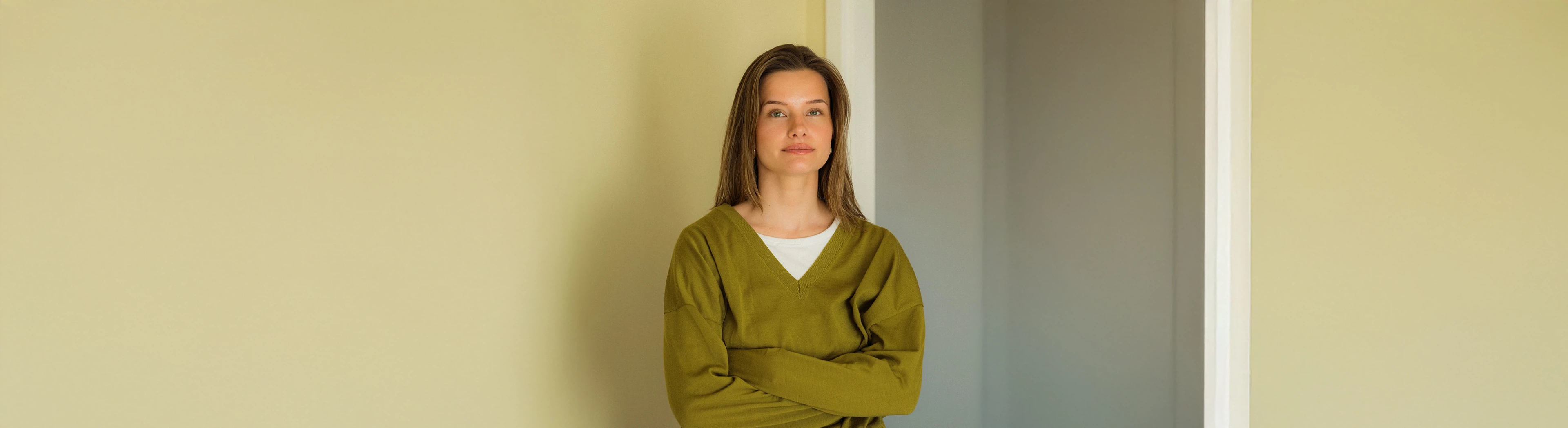 Woman with long brown hair in an olive-green sweater in front of a pale yellow wall.
